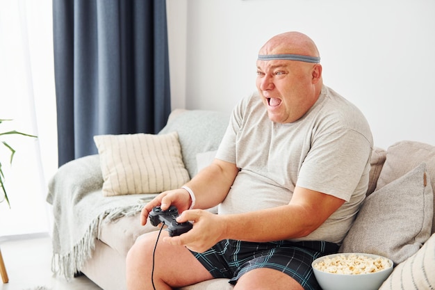 Plays video game Funny overweight man in casual clothes is indoors at home