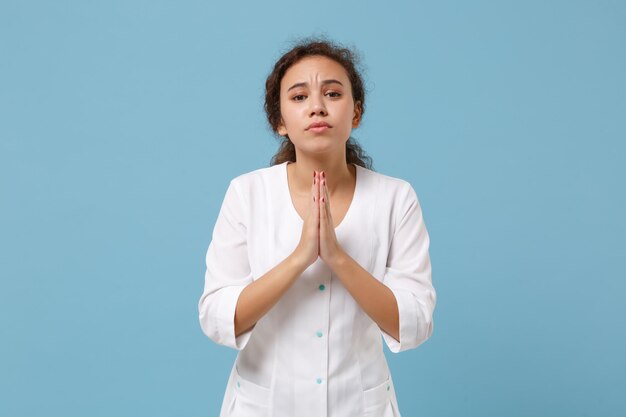 Pleading african american doctor woman isolated on blue\
background. female doctor in white medical gown hold hands folded\
in prayer. healthcare personnel health medicine concept. mock up\
copy space.