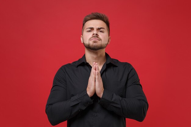 Pleading young bearded guy 20s in classic black shirt posing isolated on red background studio portrait. People sincere emotions, lifestyle concept. Mock up copy space. Holding hands folded in prayer.