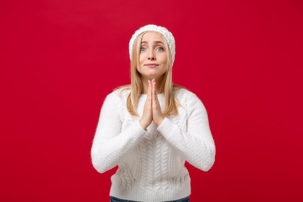 Pleading young woman in white sweater, hat isolated on red background studio portrait. Healthy fashion lifestyle, people emotions, cold season concept. Mock up copy space. Hold hands folded in prayer.