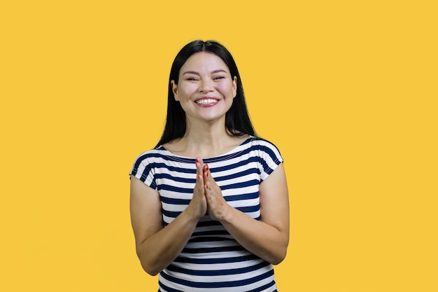 노란색 b 위에 서 있는 간청 기도 제스처에 손을 잡고 웃는 즐거운 매력적인 아시아 소녀