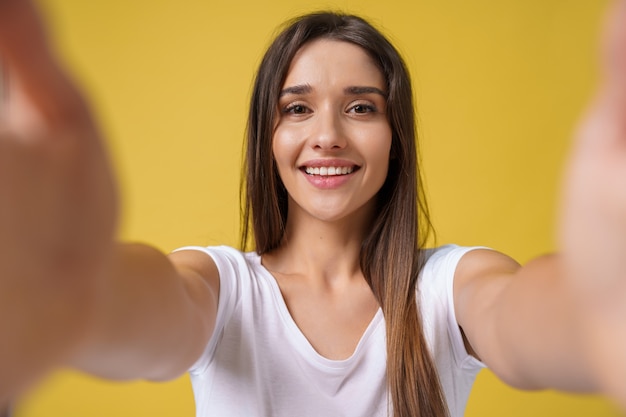 Pleasant attractive girl making selfie in studio and laughing. Good-looking young woman with brown hair taking picture of herself on bright yellow background.