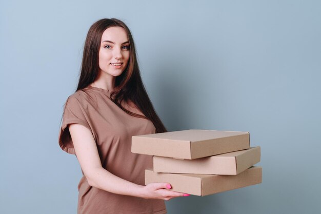 A pleasant beautiful young woman stands halfsided holding a stack of boxes on her outstretched arms on an empty blue wall