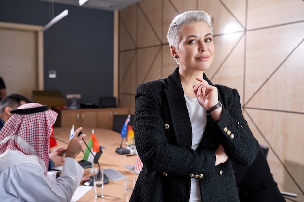 Piacevole imprenditrice con i capelli corti in abbigliamento formale in posa nella sala del consiglio durante la riunione con un gruppo internazionale di politici seduti alla scrivania