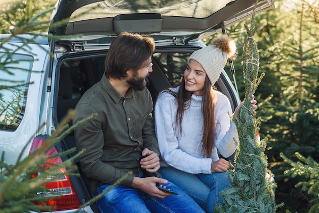 Pleasant couple sitting in car trunk and looking at wonderful christmas tree they choose for holidays