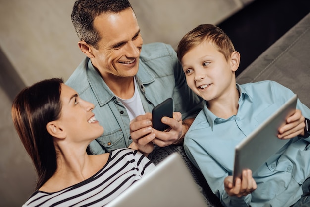 Pleasant discussion. Upbeat young family talking to each other while sitting on the sofa and using their phones