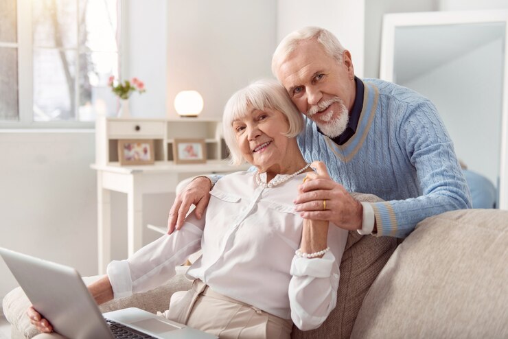 Photo pleasant distraction. joyful elderly man hugging his beloved wife from behind while she sitting on the couch and working on the laptop