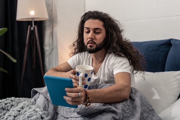 Pleasant entertainment. Nice brunette man sitting with a cup of tea while watching a video online