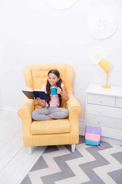 Pleasant evening with book. Girl child sit yellow armchair read book. Kid prepare to go to bed. Pleasant time cozy interior. Girl kid long hair cute pajamas relax and read fairytale book before sleep.