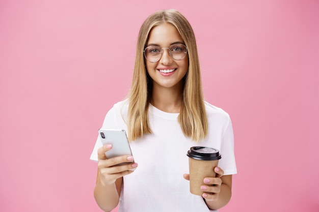pleasant friendly-looking girl smiling at camera holding paper cup of coffee and smartphone. portrait of joyful nice woman drinking morning drink, posing opinion about cafe in internet over pink wall