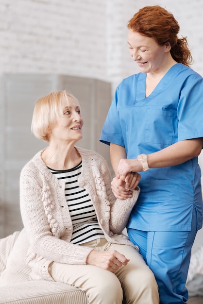 Photo pleasant helper. lovely private medical worker holding her patient by the wrist and estimating the heart rate while conducting a general checkups at ladies apartment