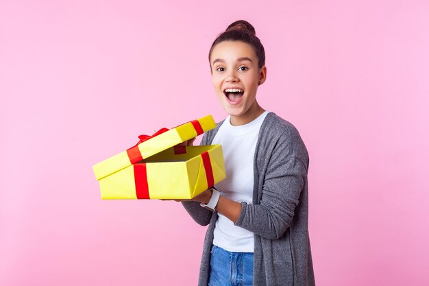 Pleasant holiday surprise. Portrait of excited happy teenage girl with bun hairstyle unboxing birthday present, screaming with joy, amazed expression. indoor studio shot isolated on pink background