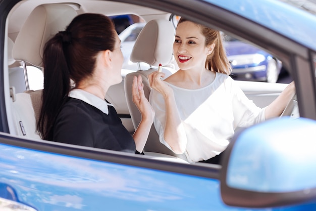 Pleasant interaction. Happy positive pretty woman sitting behind the wheel and holding a lipstick while talking to her friend