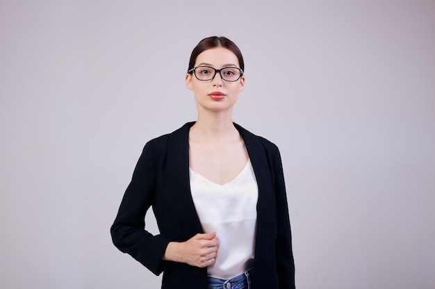 Pleasant-looking business woman is standing on gray in a black jacket, white T-shirt and computer glasses.