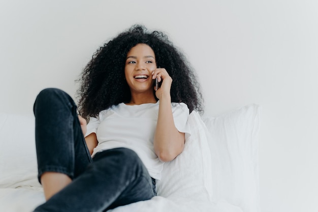 Pleasant looking curly woman has pleasant conversation talks\
with friend via smartphone while relaxes in bed has joyful\
expression looks aside wears casual clothing people bedding\
technology