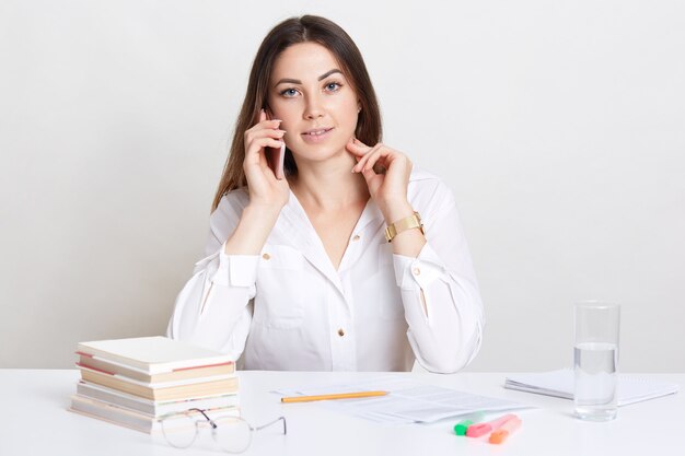 Pleasant looking dark haired woman has conversation via cell phone, involved in working process, writes some information, studies documentation, isolated over white wall. Technology concept