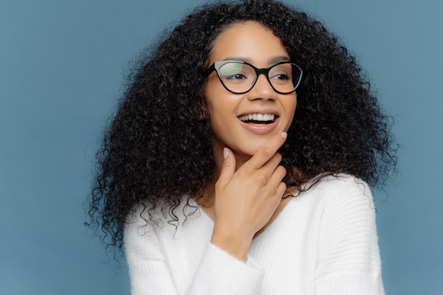 Pleasant looking dark skinned young woman holds chin, looks away, has curly hair