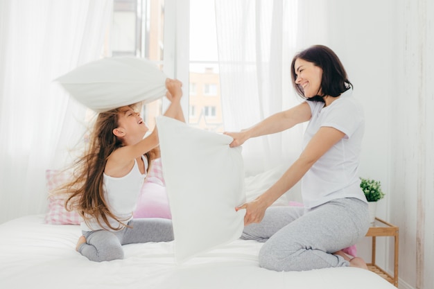 Pleasant looking female has joy together with her small daughter, have pillow fight in bedroom, pose in cozy spacious light room