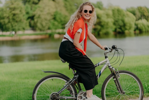 Pleasant looking satisfied curly haired woman spends free time in open air rides bicycle spends leisure alone enjoys freshness of nature looks at camera with pleasant smile Summer time