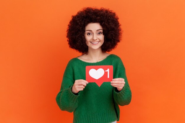 Pleasant looking woman with Afro hairstyle wearing green sweater standing with social media heart Like icon, recommending to follow and love content. Indoor studio shot isolated on orange background.