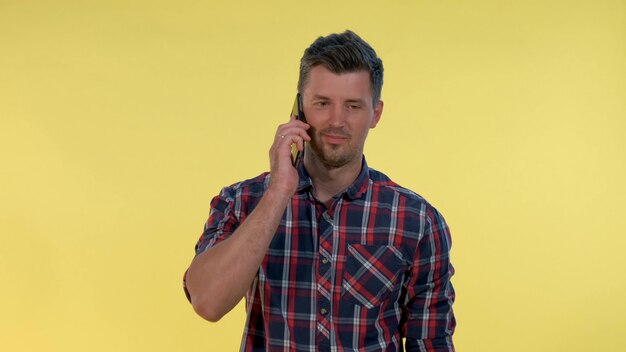 Pleasant-looking young man speaking with somebody by smartphone on yellow background.