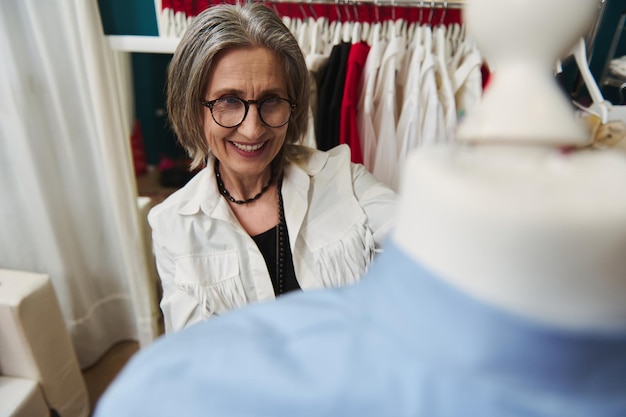 Pleasant mature female fashion designer in her tailoring\
atelier preparing new collection of exclusive clothes for\
exhibition