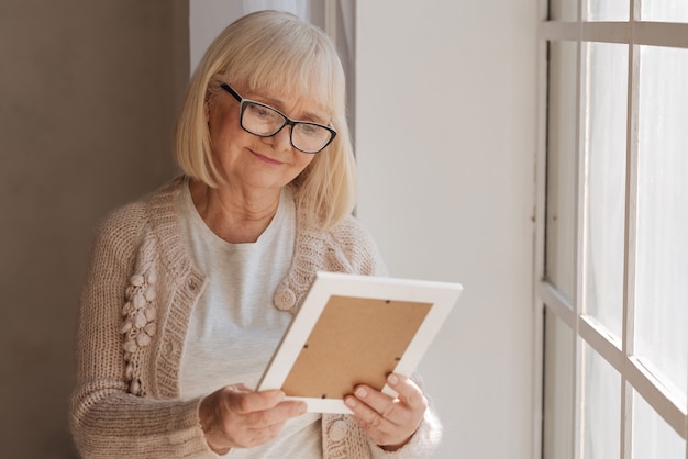 Pleasant memories. Nice positive grey haired woman looking at the photograph and smiling while standing near the window