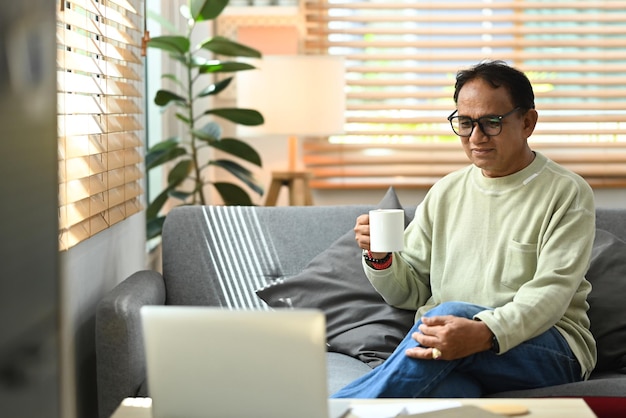 Pleasant middle aged man drinking coffee and reading online news or browsing internet on laptop at home