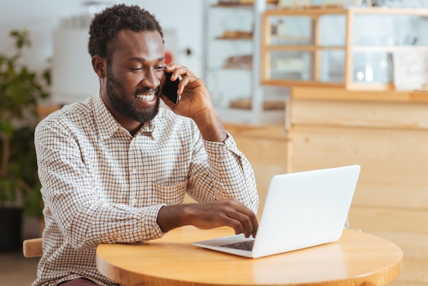 Pleasant news. Joyful handsome man sitting at the table in the coffee house, receiving good news about work on the phone while typing on laptop with one hand