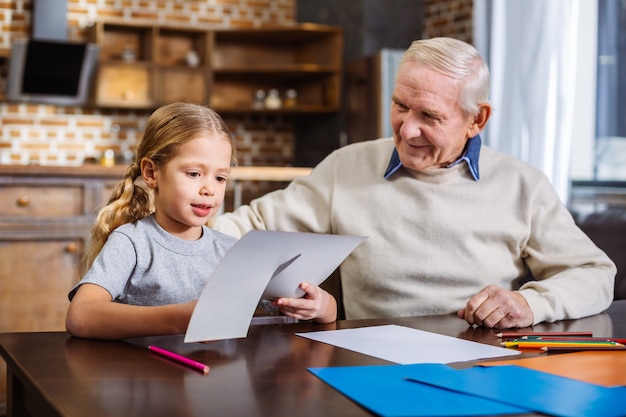 Pleasant nice little girl cutiting the paper while sitting at the table with her grandfather