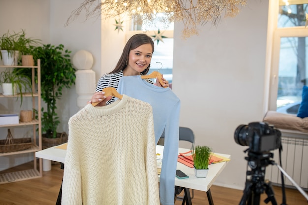 Pleasant nice woman standing in front of the camera while holding two pieces of clothes