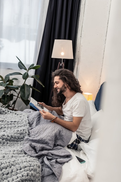 Pleasant pastime. Joyful bearded man reading a book while relaxing at home