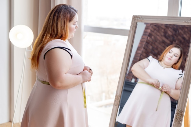 Приятная пухленькая женщина смотрит в зеркало, измеряя талию