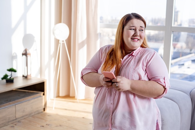 Pleasant plump woman typing a message on her smartphone while standing in the living room