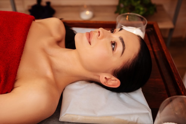 Pleasant procedure. Calm good-looking woman with long hair peacefully lying naked on wooden table covered with red towel