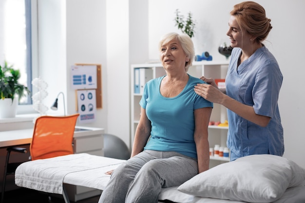 Pleasant procedure. Positive aged woman smiling while receiving professional massage