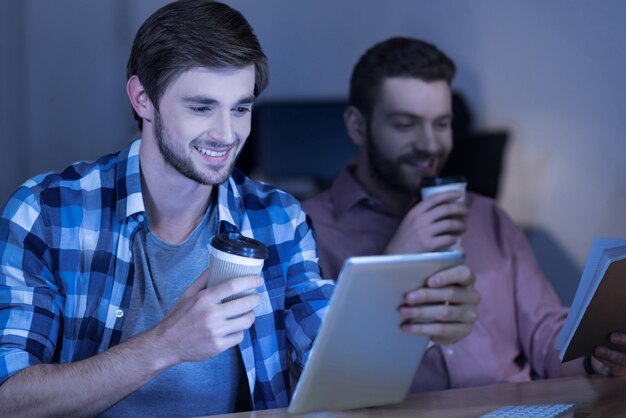 Pleasant rest. Happy delighted handsome man reading something on the tablet screen and smiling while having coffee