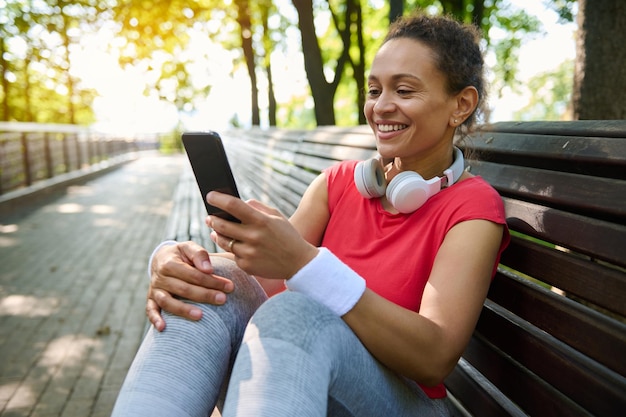 Pleasant smiling african american sportswoman athlete with\
headphones and white terry wristbands using smartphone checks\
fitness mobile app after workout outdoors relaxes on wooden bench\
in city park