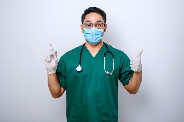 Pleasant smiling asian male doctor physician in medical mask and gloves show thumbup hold syringe with vaccine