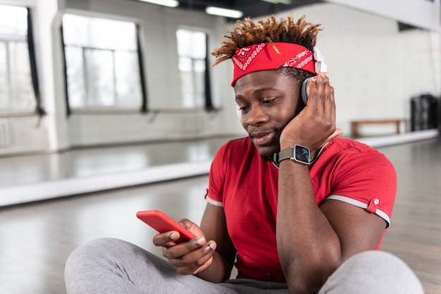 Pleasant smiling short haired guy listening music through headphones while sitting on floor