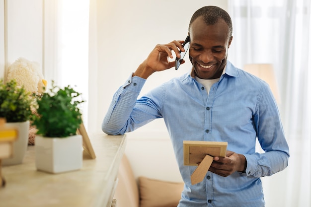 Pleasant talk. Handsome inspired young man talking on the phone and smiling while holding and looking at the photo