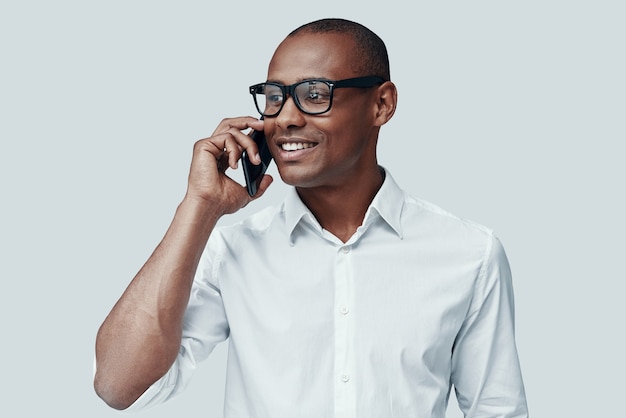 Pleasant talk. Handsome young African man talking on the smart phone while standing against grey background