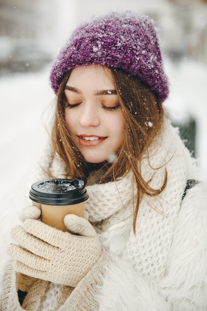 Pleasant young lady with closed eyes in warm clothing holding hot coffee-to-go in hands while posing outdoors.