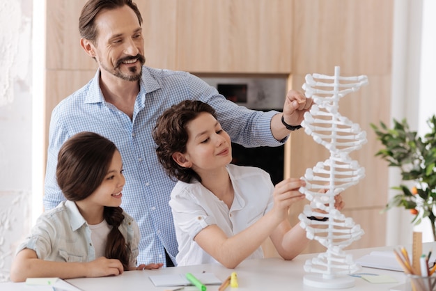 Foto piacevole giovane che punta a un grande modello di dna 3d con una matita mentre suo figlio tocca i nucleotidi e sua figlia minuta guarda il modello con un sorriso