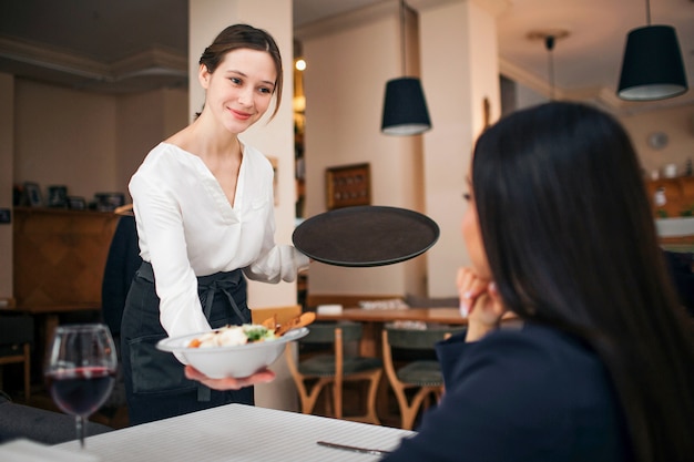 Pleasant young waitress stand at table and shows bowl of salad