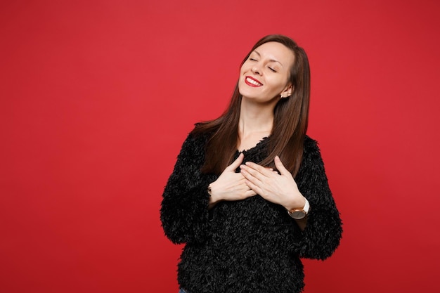 Pleasant young woman in black fur sweater keeping eyes closed, holding hands on chest, heart isolated on bright red wall background. People sincere emotions, lifestyle concept. Mock up copy space.