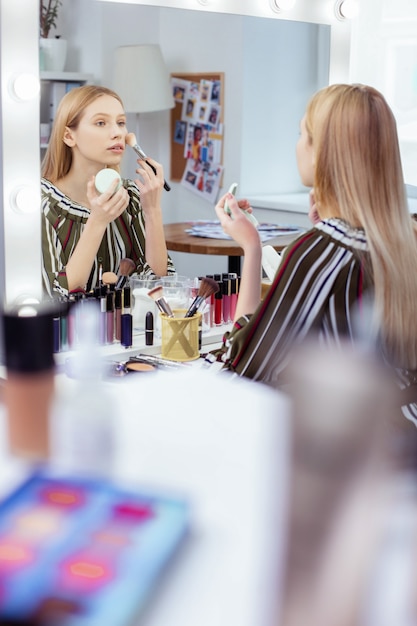 Приятная молодая женщина смотрит на себя в зеркало во время макияжа