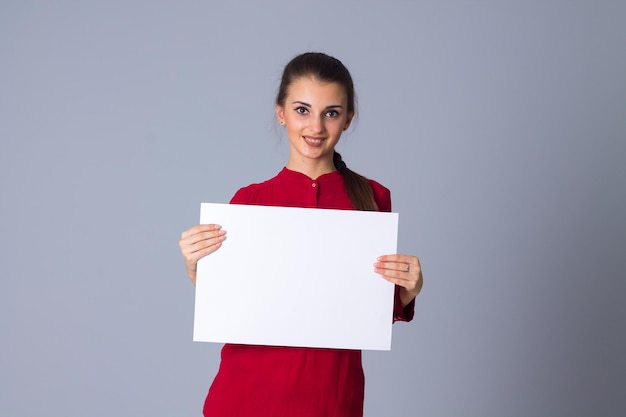 Pleasant young woman in red blouse holding white sheet of paper on grey background in studio