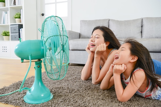 pleasantly mother with young little daughter lying down on living room floor and face to electric fan enjoying blowing cool wind together at summer.