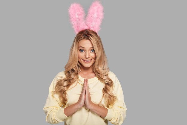 Please! Attractive young woman in pink bunny ears keeping hands clasped and looking at camera while standing against grey background